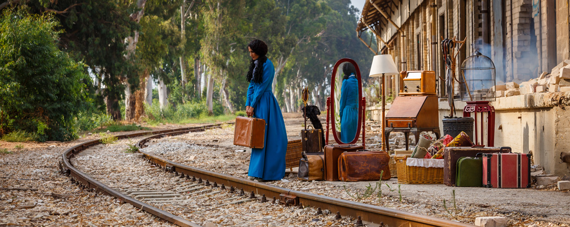 Image of a woman standing by railway tracks with a suitcase in her hands.